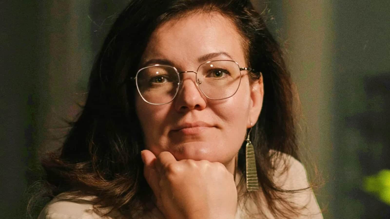 Woman with glasses and long brown hair rests her chin on her hand, looking at the camera. She wears a beige top, dangling earrings, and appears thoughtful. The background is softly blurred.