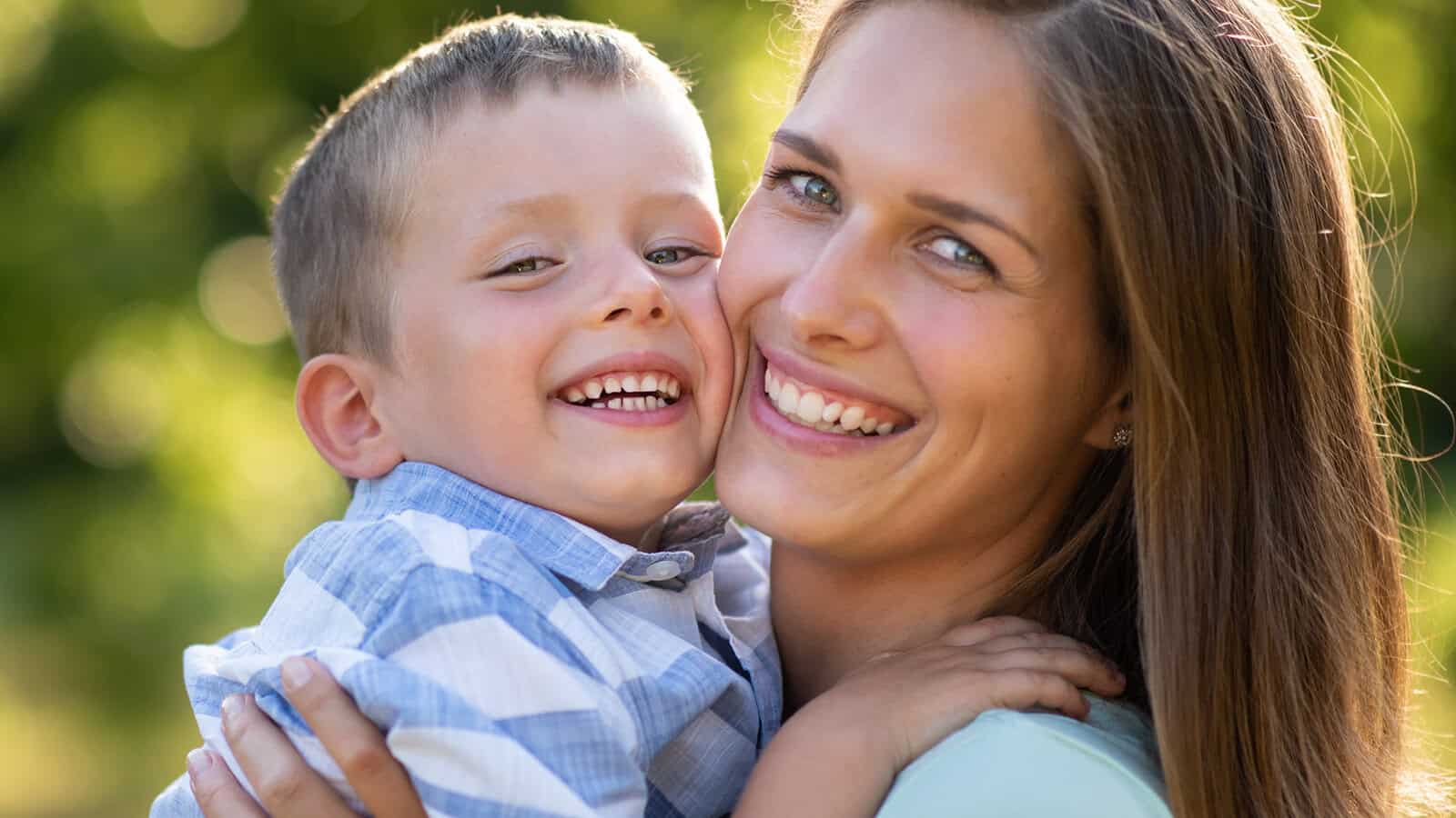 A smiling woman and a young boy hug each other closely outdoors, both looking at the camera with big smiles. Sunlight gently highlights their faces, with a blurred green background behind them.