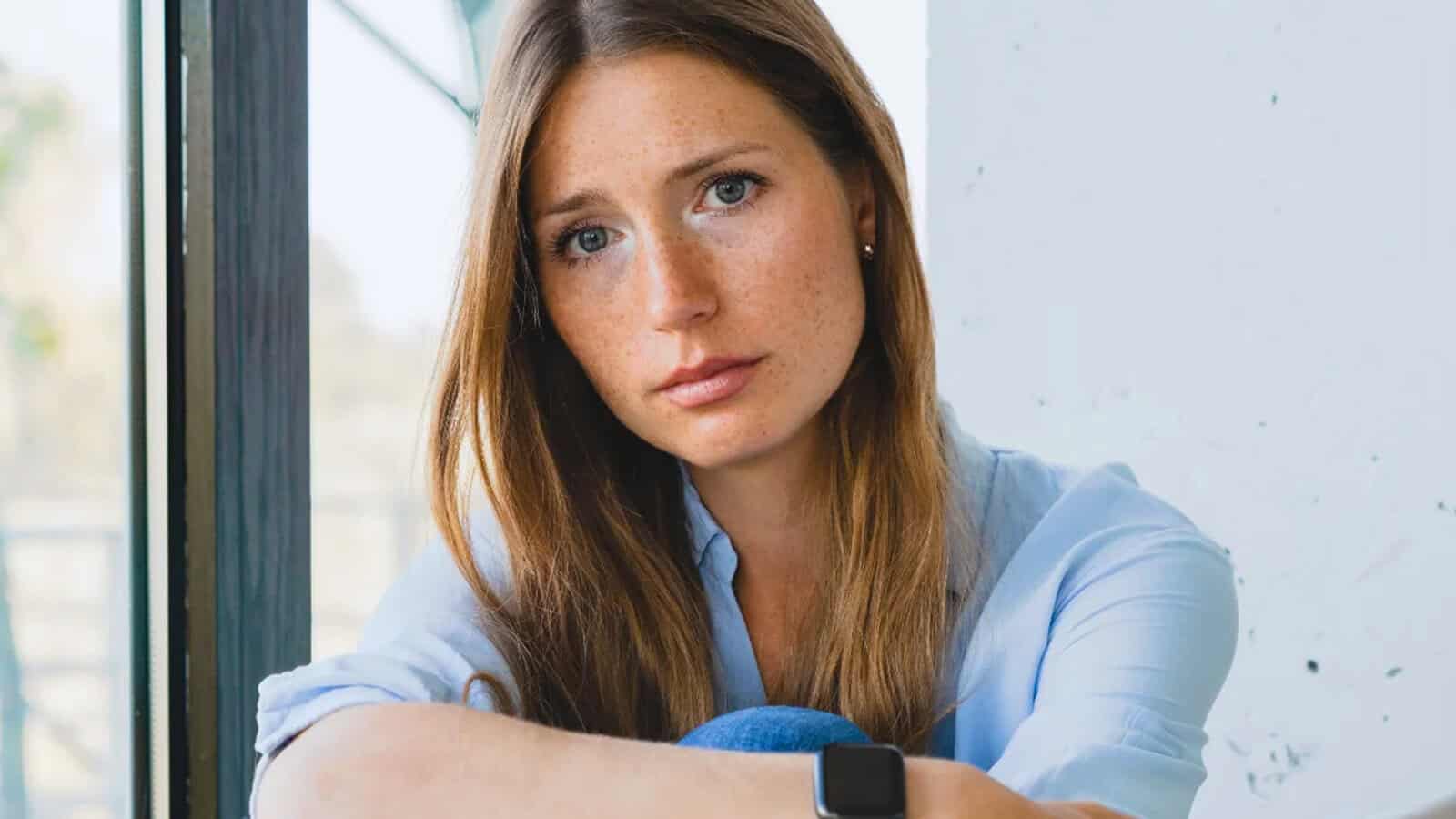 A woman with long brown hair and freckles sits indoors by a window, looking thoughtfully at the camera. She is wearing a light blue shirt and a smartwatch, with natural light illuminating her face.