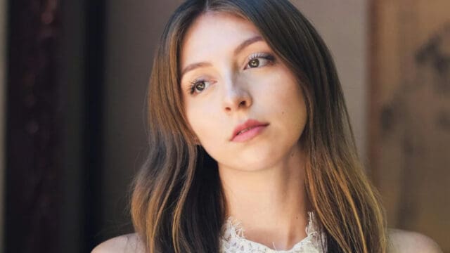A young woman with long brown hair gazes thoughtfully into the distance. She wears a sleeveless white lace top and stands in soft natural light with a neutral background.