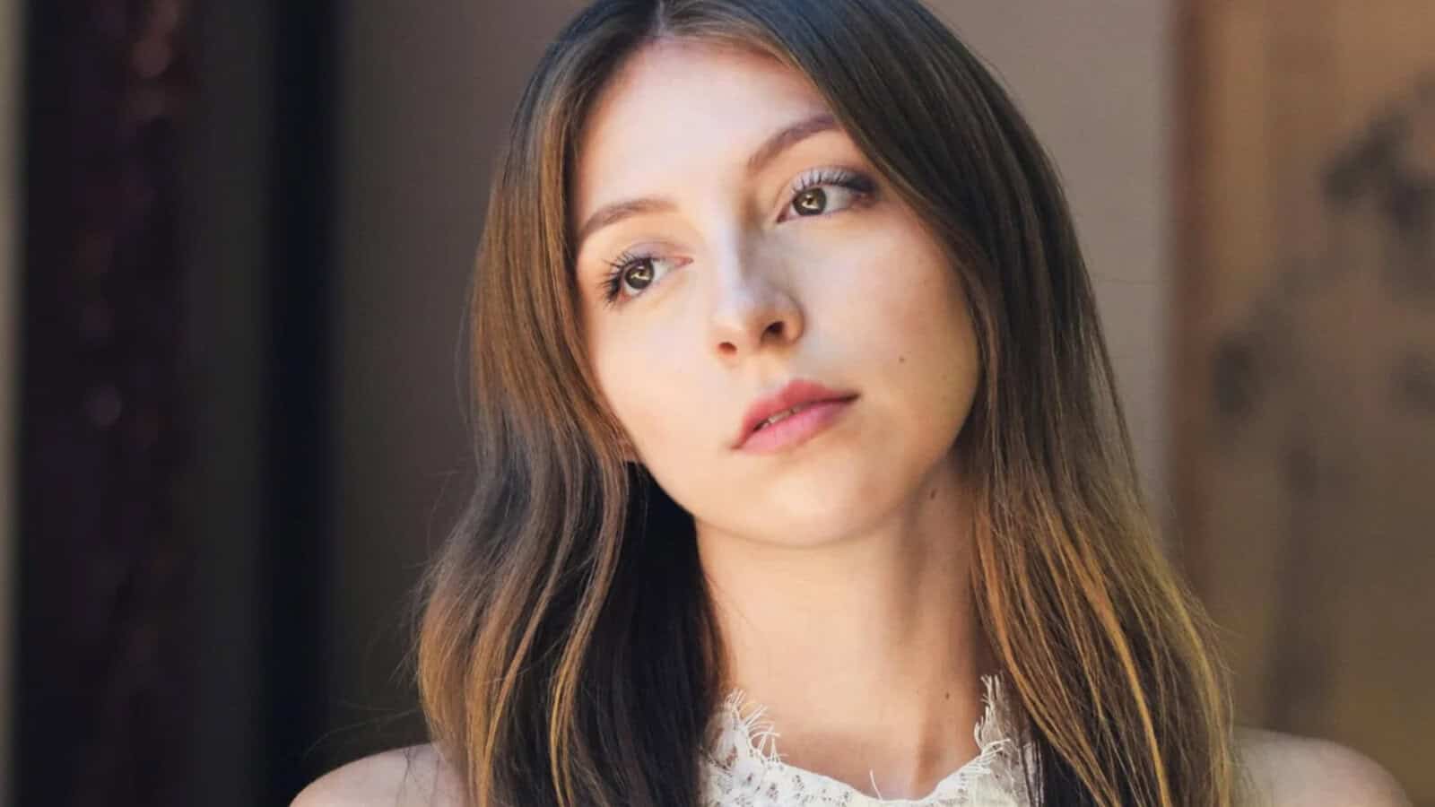 A young woman with long brown hair gazes thoughtfully into the distance. She wears a sleeveless white lace top and stands in soft natural light with a neutral background.