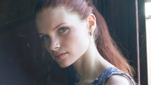 A woman with long reddish-brown hair, wearing a patterned top, looks seriously at the camera while standing near a wooden door, illuminated by soft natural light.
