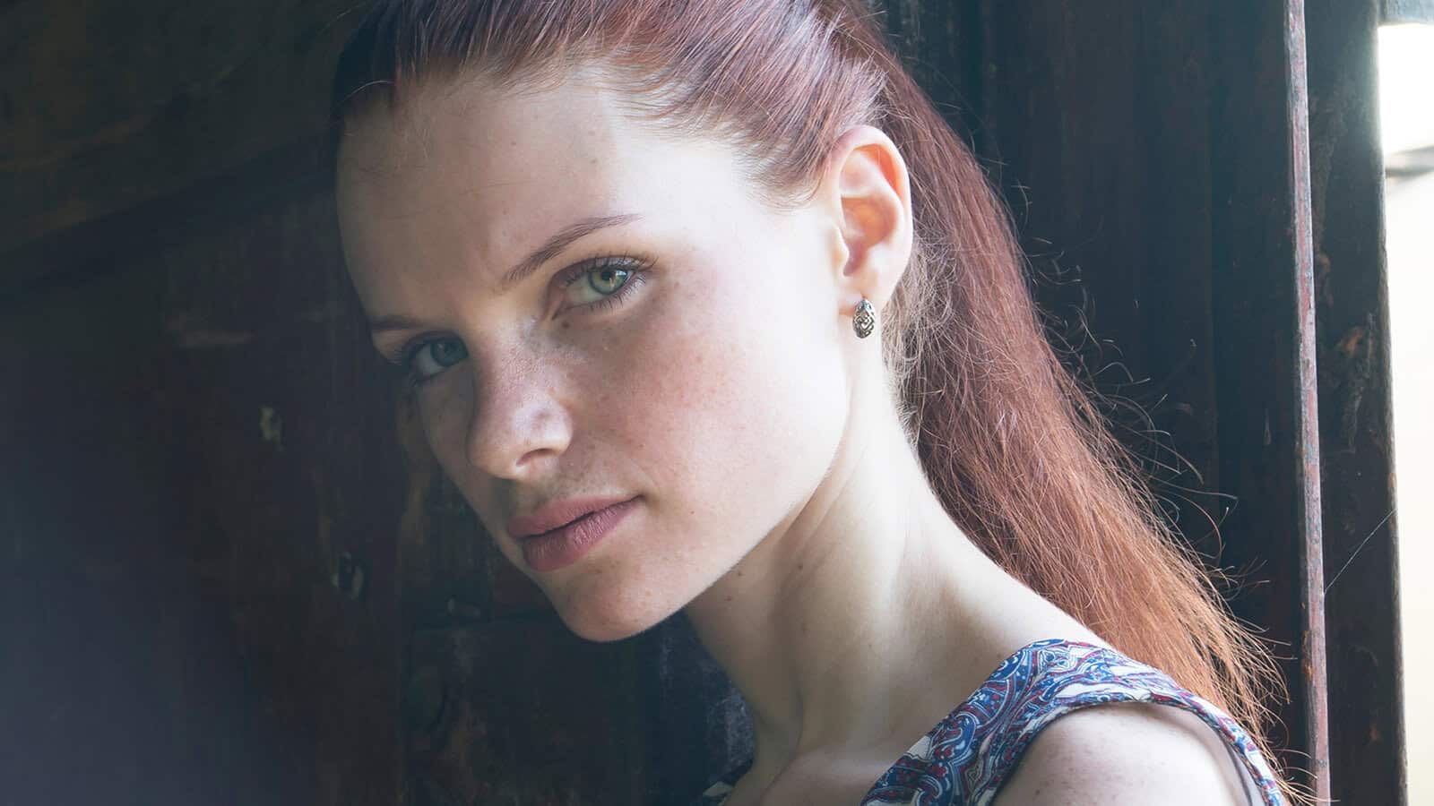 A woman with long reddish-brown hair, wearing a patterned top, looks seriously at the camera while standing near a wooden door, illuminated by soft natural light.
