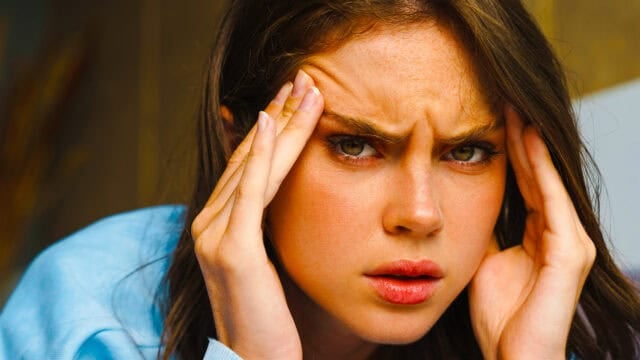 A woman with long brown hair looks directly ahead, furrowing her brows and holding her hands to her temples, appearing stressed or frustrated.