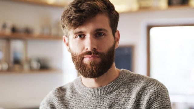 A man with a brown beard and short hair wearing a gray knit sweater looks at the camera in a softly lit indoor setting with blurred shelves in the background.