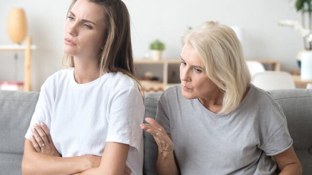 Younger woman sits with arms crossed, looking away, while older woman beside her gestures with concern, appearing to speak to her. Both women look serious, suggesting a disagreement or difficult conversation.
