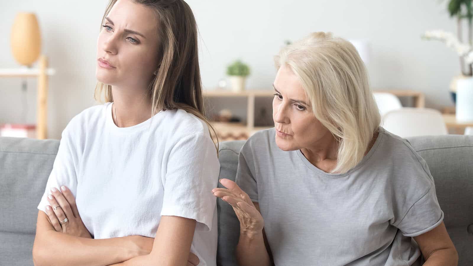 Younger woman sits with arms crossed, looking away, while older woman beside her gestures with concern, appearing to speak to her. Both women look serious, suggesting a disagreement or difficult conversation.