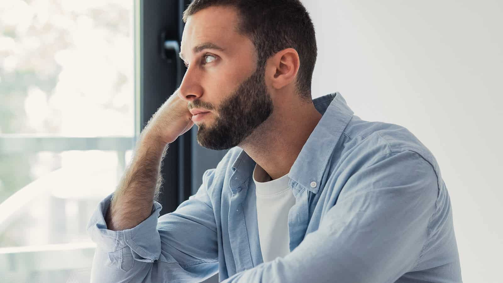 A man with a beard, wearing a light blue shirt, sits indoors by a window, resting his head on his hand and looking thoughtfully into the distance.