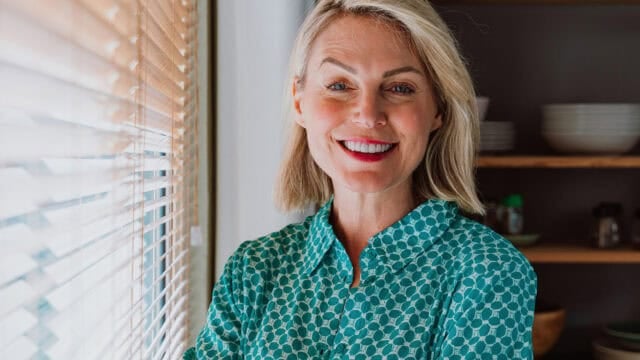 Smiling woman with blonde hair wearing a teal patterned shirt stands by a window with blinds, with shelves and dishes visible in the background.