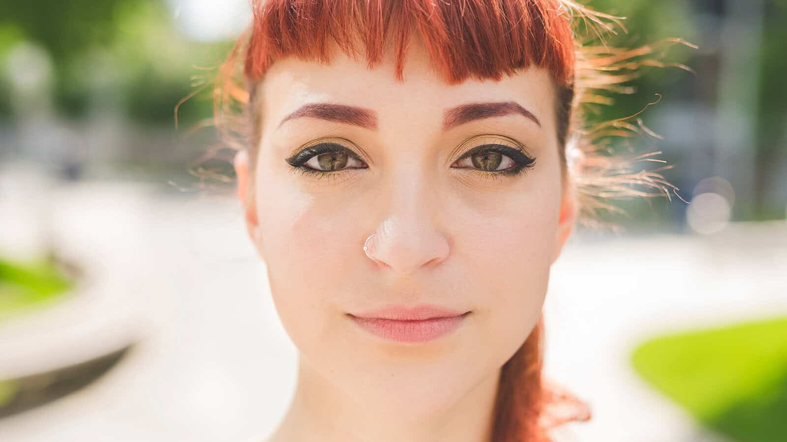 A person with red hair and winged eyeliner looks into the camera outdoors on a sunny day, with blurred green trees and a pathway in the background.