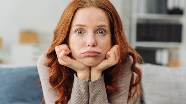 A woman with long red hair rests her chin on her hands, cheeks puffed out, and looks directly at the camera with a frustrated or bored expression.