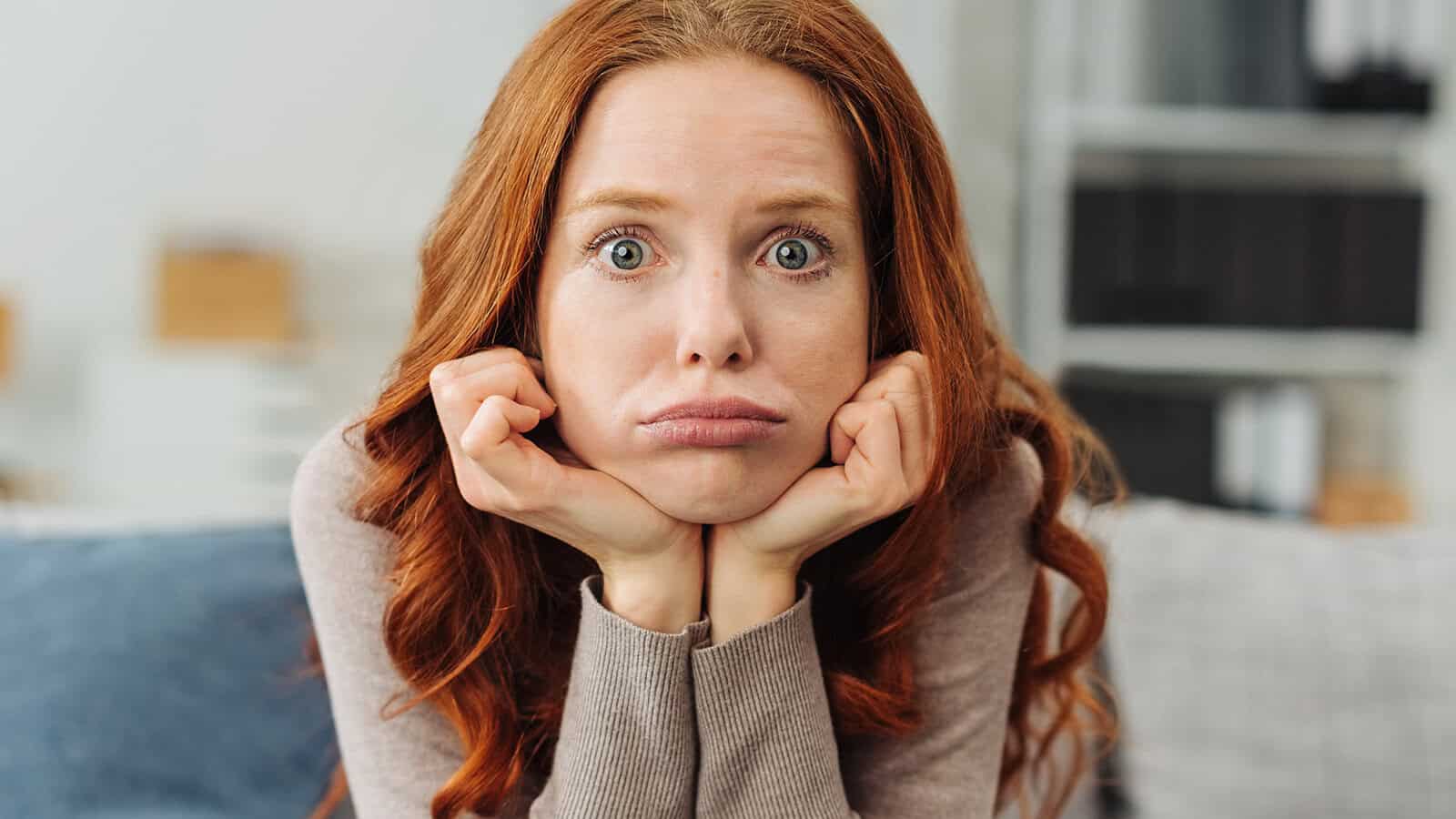 A woman with long red hair rests her chin on her hands, cheeks puffed out, and looks directly at the camera with a frustrated or bored expression.