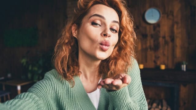 A woman with curly red hair wearing a green sweater blows a kiss toward the camera while indoors. A wooden wall and a clock are visible in the background.