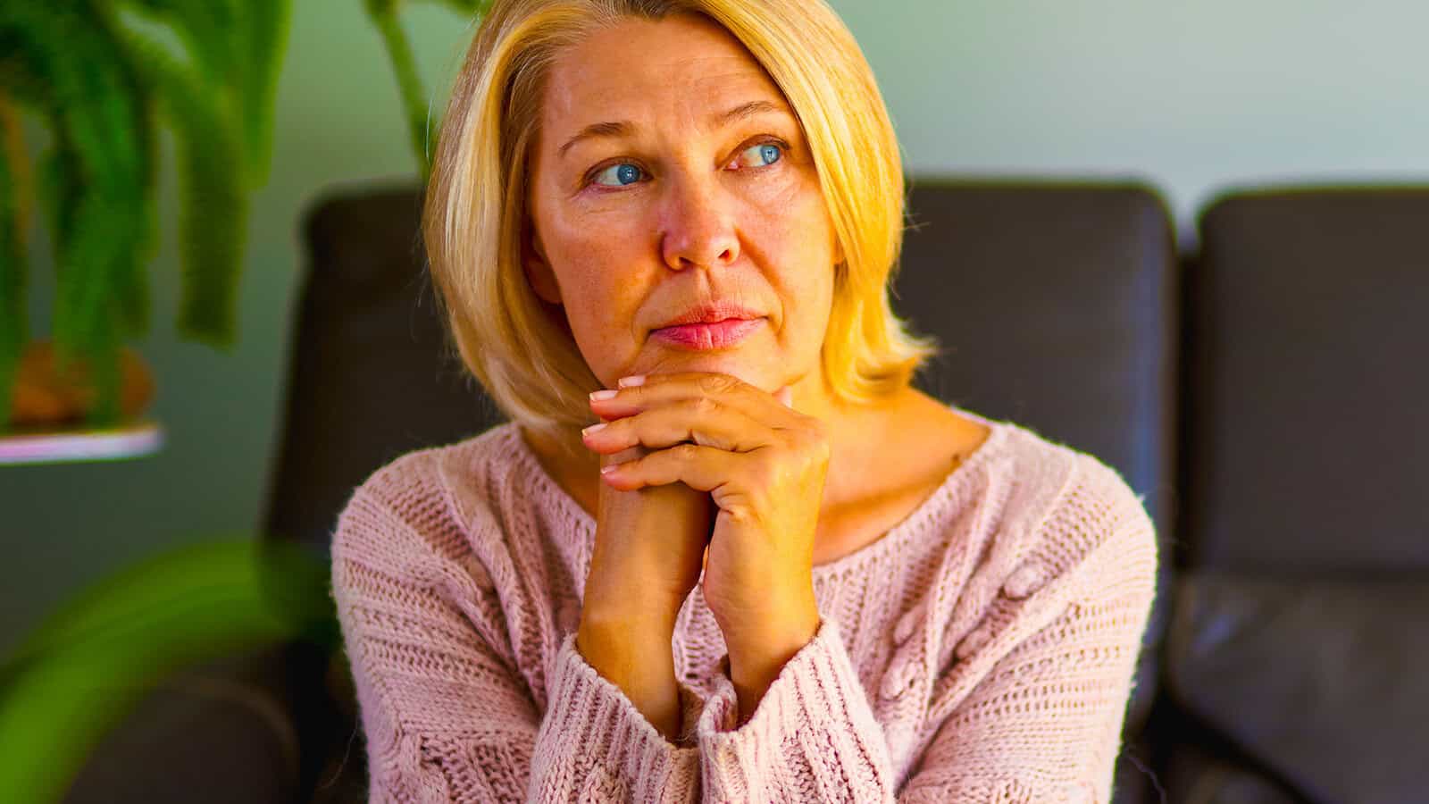 A woman with short blonde hair in a pink sweater sits indoors, resting her chin on her hands and gazing thoughtfully to the side. A plant is visible in the background.