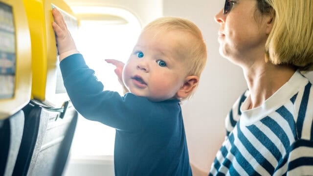 A baby with blonde hair looks over their shoulder while standing on an airplane seat next to an adult in sunglasses and a striped shirt.