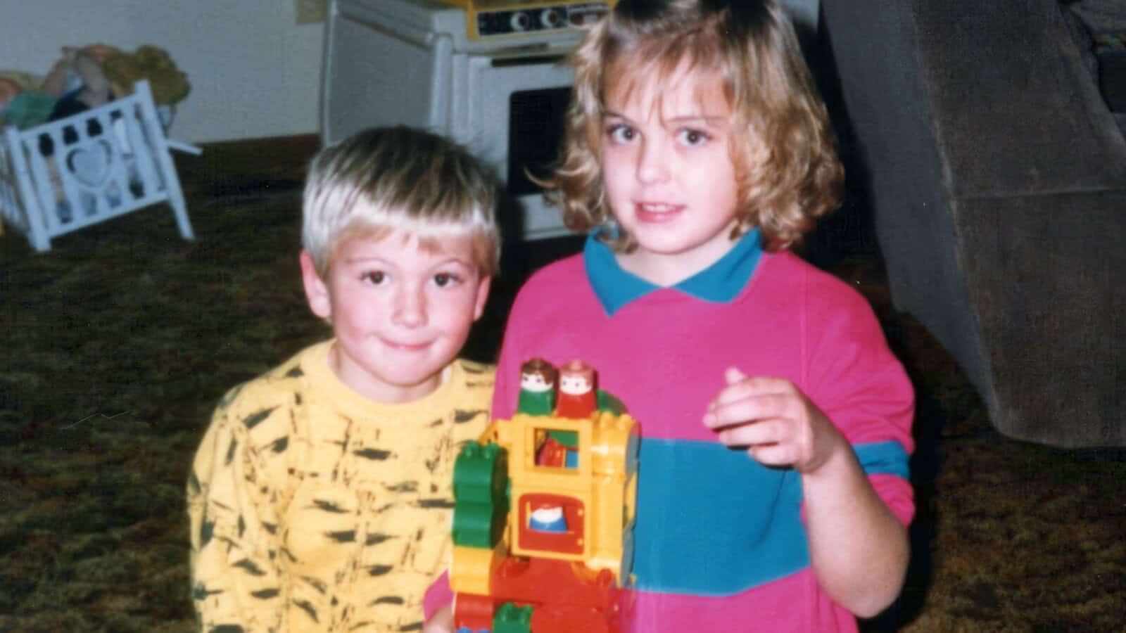 Two young children stand close together indoors, smiling and holding a colorful toy made of interlocking plastic blocks. The background shows part of a kitchen and some furniture.