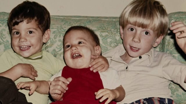 Three young children sit on a green patterned couch, smiling and laughing together. The child in the center wears a red outfit, while the other two wear light-colored shirts. They appear happy and playful.