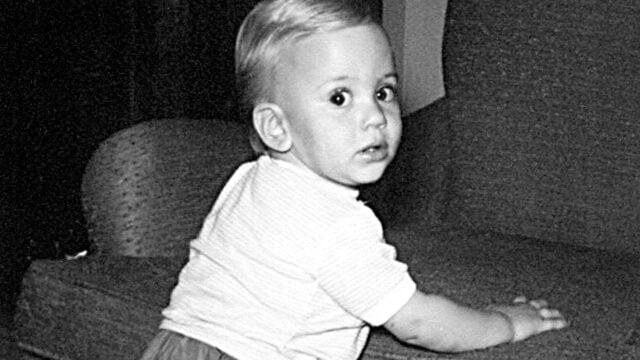 A black-and-white photo of a baby with light hair and a striped shirt, standing and holding onto a couch, looking over their shoulder toward the camera.