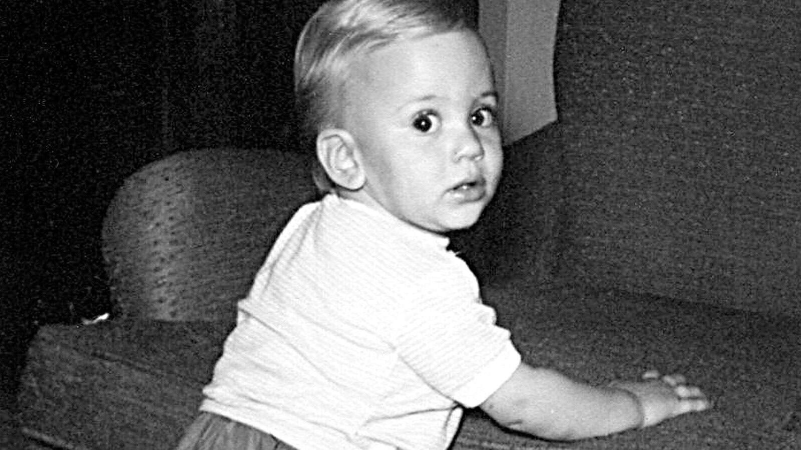 A black-and-white photo of a baby with light hair and a striped shirt, standing and holding onto a couch, looking over their shoulder toward the camera.