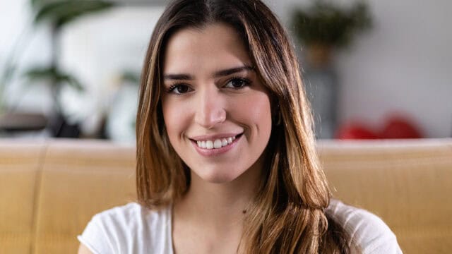 A young woman with long brown hair and a white shirt smiles while sitting indoors on a yellow couch, with blurred plants in the background.