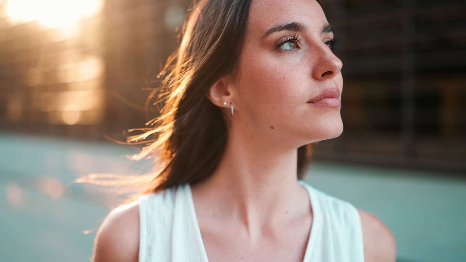 A young woman with long brown hair looks thoughtfully into the distance, lit by soft sunlight. She wears a sleeveless light top and stands outdoors, with a blurred background behind her.