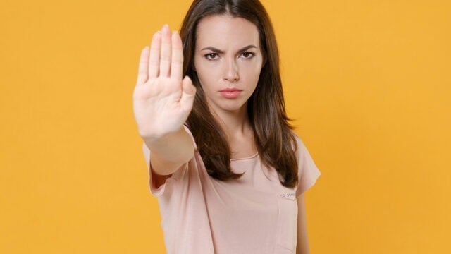 A woman in a light pink shirt stands against a yellow background, holding her hand up with her palm facing forward in a stop gesture, looking serious.