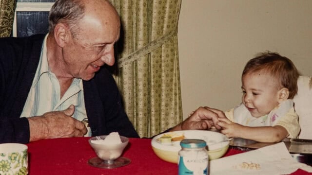 An elderly man smiles at a toddler sitting at a table with food, sharing a happy moment. The table has a red tablecloth, and there are jars, a bowl, and a mug visible.