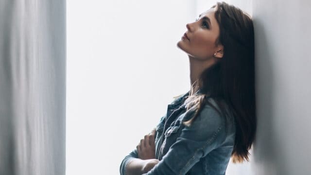 A woman with long brown hair and a denim jacket stands in a narrow hallway, leaning back against the wall, looking upwards with her arms folded across her chest. Light shines from one side, creating a soft, contemplative mood.