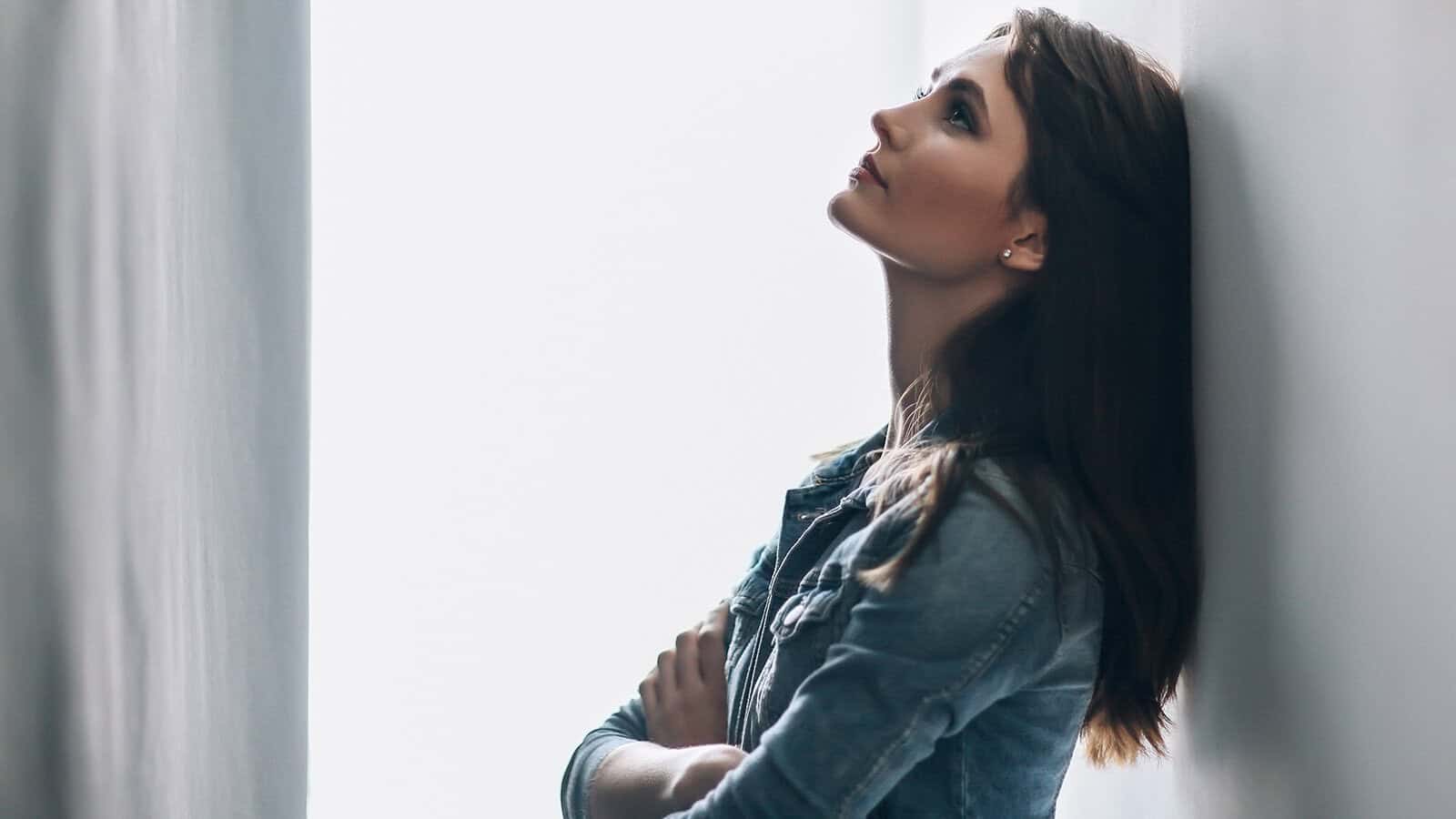 A woman with long brown hair and a denim jacket stands in a narrow hallway, leaning back against the wall, looking upwards with her arms folded across her chest. Light shines from one side, creating a soft, contemplative mood.