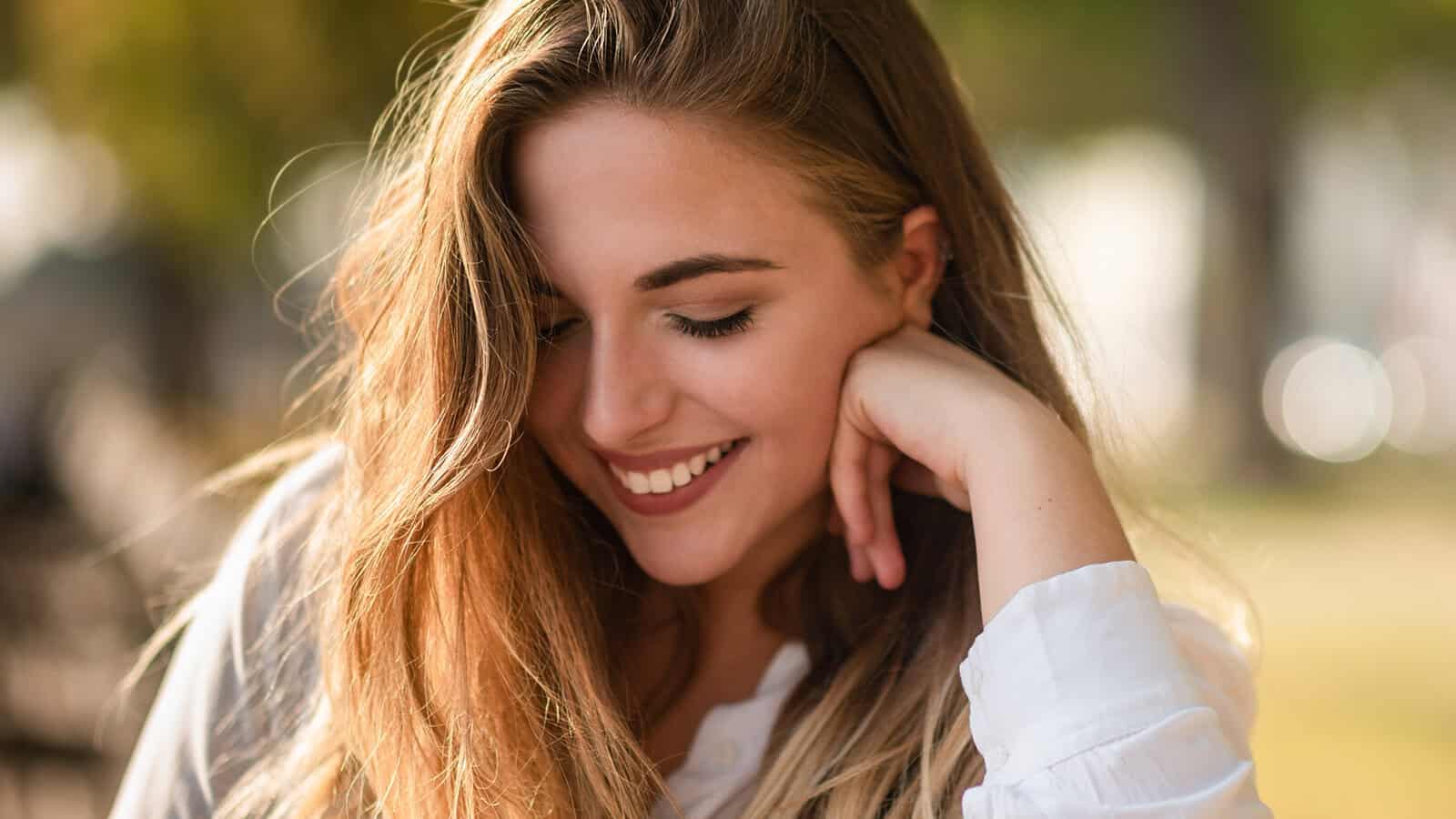 A young woman with long blonde hair smiles softly while looking down, resting her chin on her hand, outdoors with blurred trees in the background.