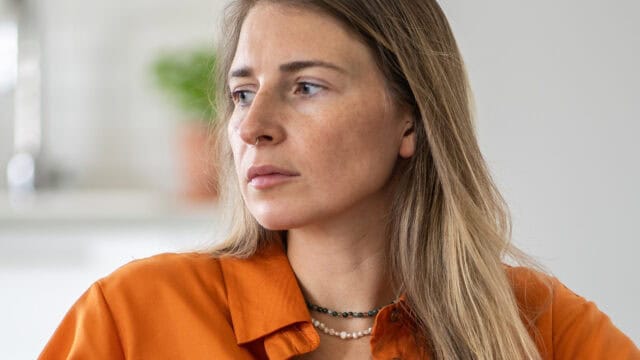 A woman with long blonde hair wearing an orange shirt looks to the side with a serious expression. She wears two beaded necklaces. The background is softly blurred.