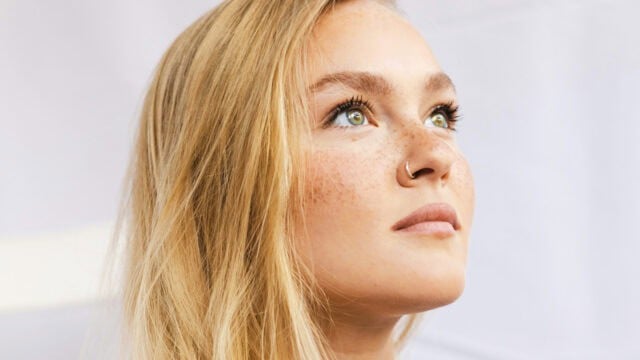A close-up of a young woman with long blonde hair, light eyes, freckles, and a nose ring, looking upward with a thoughtful expression. The background is softly blurred and light-colored.