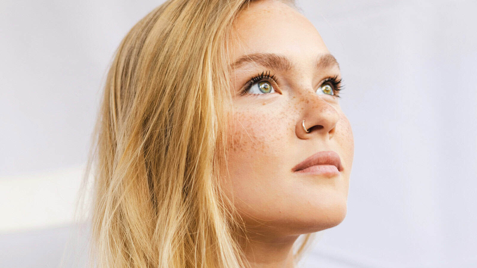 A close-up of a young woman with long blonde hair, light eyes, freckles, and a nose ring, looking upward with a thoughtful expression. The background is softly blurred and light-colored.