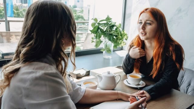 Two women sit at a table with tea, having a conversation. One gestures while speaking. There is a plant and window in the background, and a notepad and pen on the table.