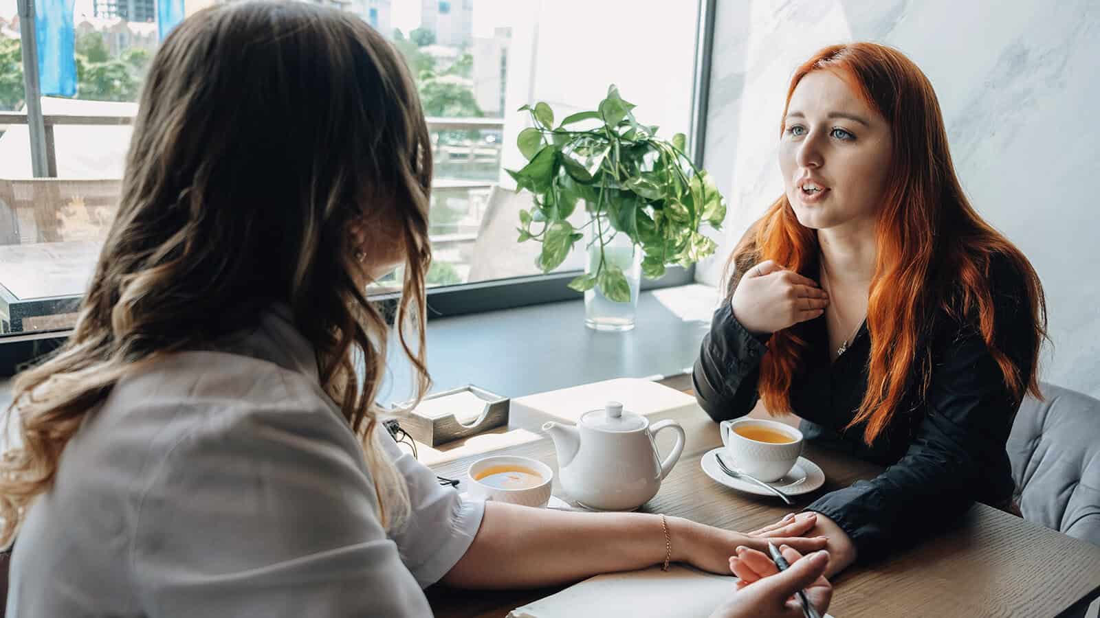 Two women sit at a table with tea, having a conversation. One gestures while speaking. There is a plant and window in the background, and a notepad and pen on the table.