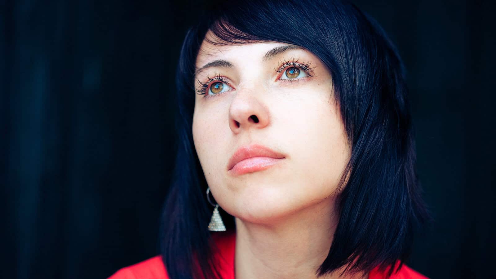 A person with short dark hair and light eyes looks upward with a thoughtful expression, wearing a red top and dangling earrings, against a dark background.
