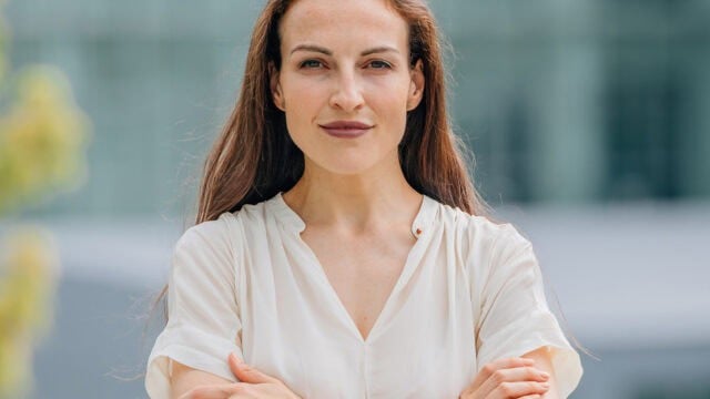 A woman with long brown hair and a white blouse stands outside with her arms crossed, looking confidently at the camera. The background is blurred and appears to be a modern building.