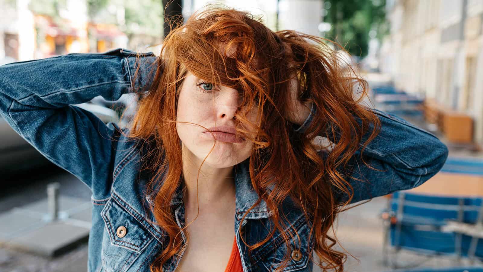 A person with long, wavy red hair and a denim jacket poses outside, hands in their hair, with strands falling across their face. The background shows blurred outdoor seating and buildings.