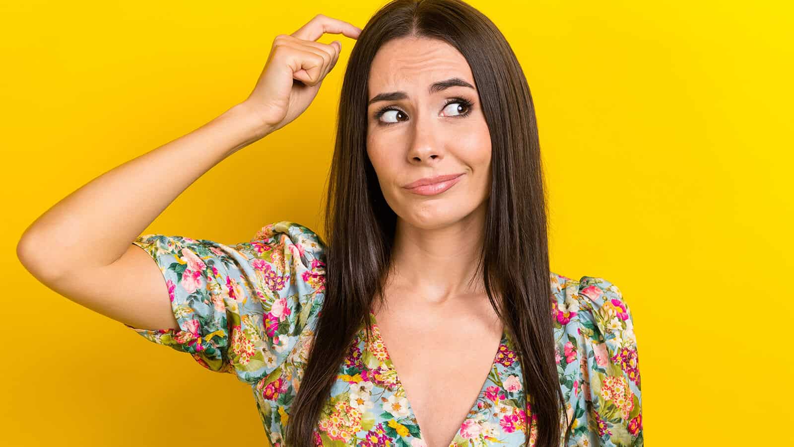 A woman with long dark hair in a floral dress scratches her head and looks confused, standing against a bright yellow background.