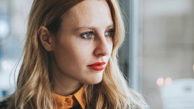 A woman with long blonde hair and red lipstick looks out a window thoughtfully. She wears a mustard yellow collared shirt and a dark sweater. Soft natural light illuminates her face.