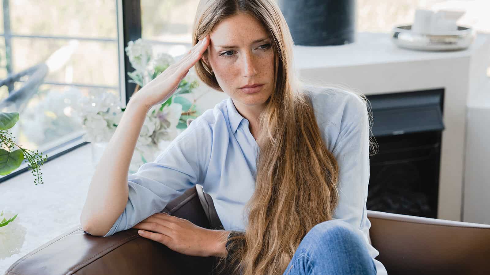 A young woman with long, light brown hair sits on a couch, resting her head on her hand and looking thoughtful or concerned. She wears a light blue shirt and jeans. The background shows flowers and a window.