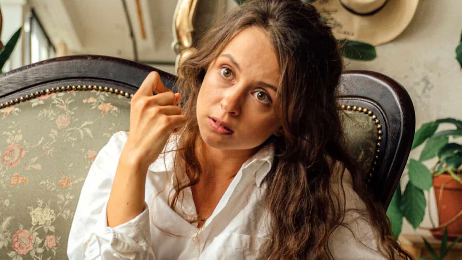 A woman with long brown hair sits in an ornate chair, wearing a white shirt. She looks intently forward with a serious expression, holding one hand near her face. There are plants and a hat in the background.