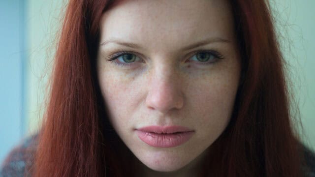 A woman with long red hair and green eyes looks directly at the camera with a neutral expression. The image is a close-up portrait, showing her freckles and natural makeup.