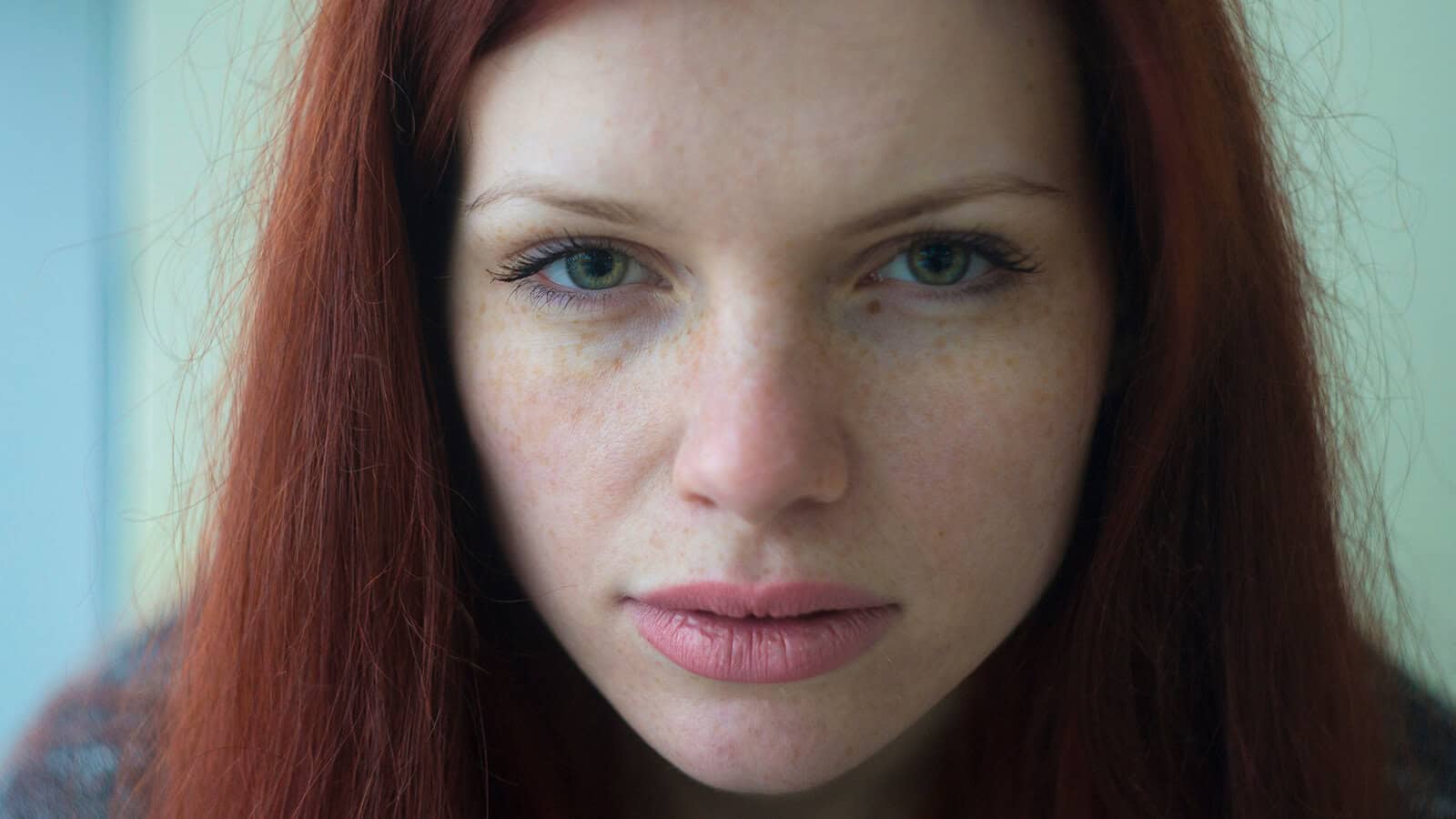 A woman with long red hair and green eyes looks directly at the camera with a neutral expression. The image is a close-up portrait, showing her freckles and natural makeup.