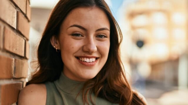 A young woman with long brown hair and a green sleeveless top smiles while leaning against a brick wall outdoors on a sunny day.