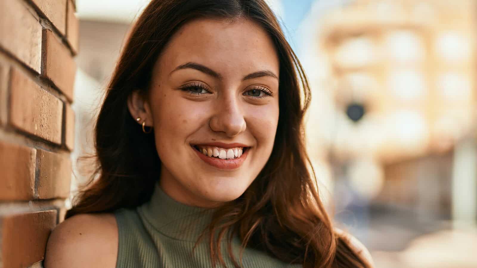 A young woman with long brown hair and a green sleeveless top smiles while leaning against a brick wall outdoors on a sunny day.