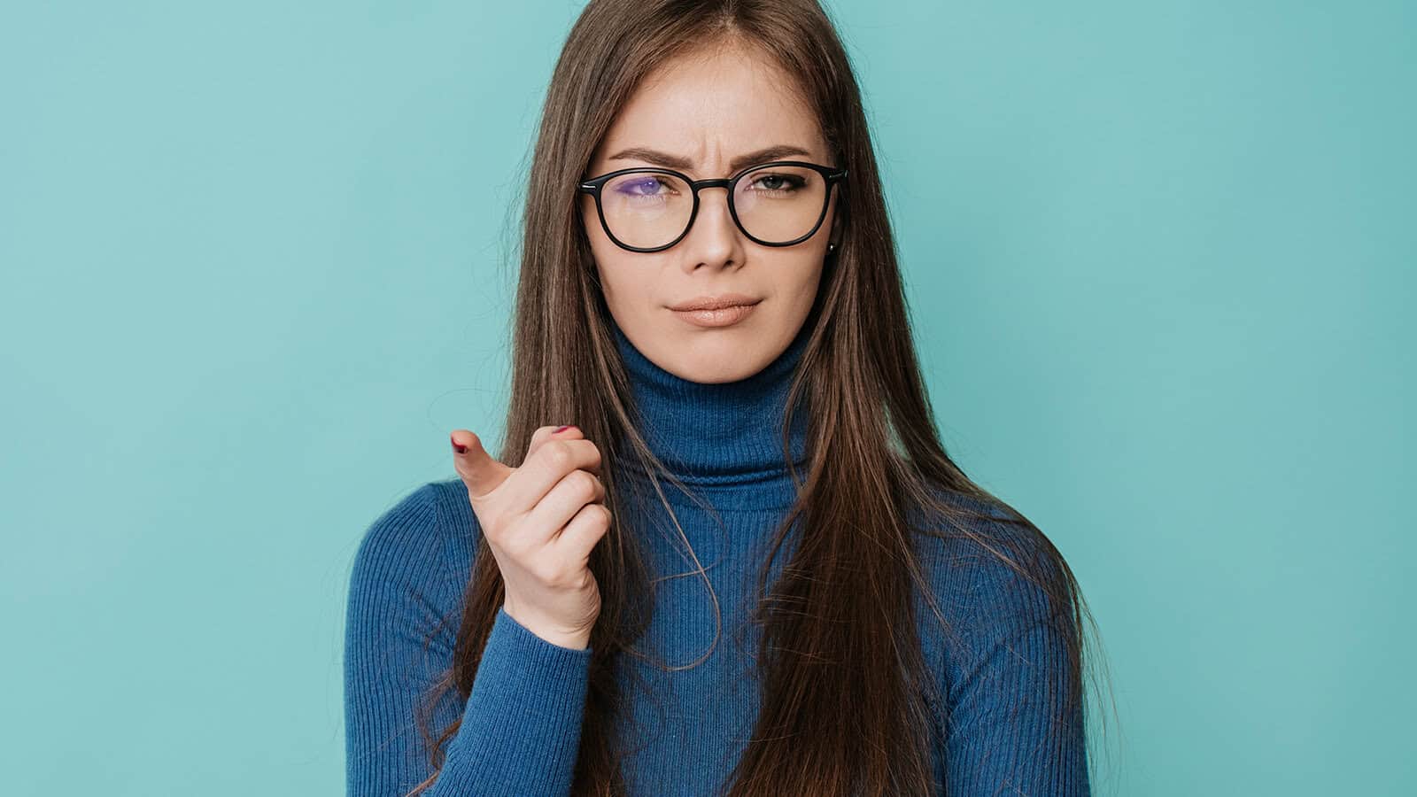 A woman with long brown hair, wearing glasses and a blue turtleneck, stands against a teal background, looking skeptical and pointing forward.