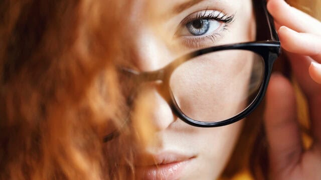 Close-up of a person with wavy red hair wearing black-framed glasses, looking directly at the camera. One hand adjusts the glasses, and one blue eye is clearly visible.