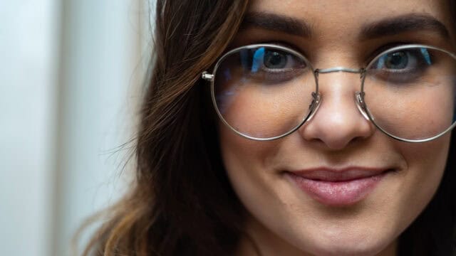 A close-up of a person with long brown hair, wearing round glasses and smiling slightly, with a blurred background.