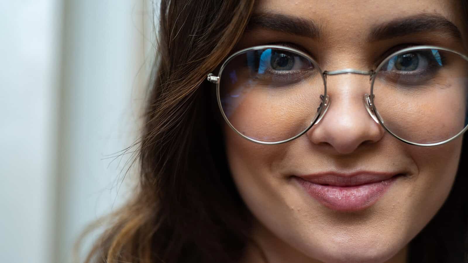 A close-up of a person with long brown hair, wearing round glasses and smiling slightly, with a blurred background.